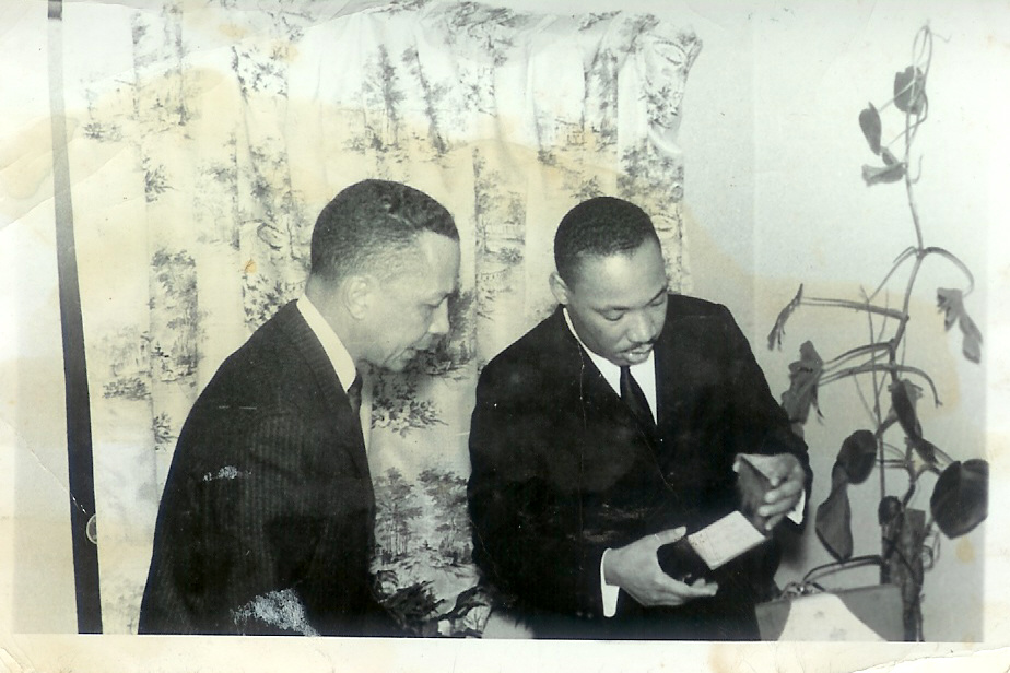 Black-and-white photograph of Dr. Rodgers and Dr. Martin Luther King Jr. standing side by side in dark suits, looking down at a small booklet Dr. King holds, in front of a patterned curtain.